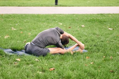 Spor giyimli genç bir adam parkta yoga yapıyor. Asana 'yı dışarıda pratik yap. Yeşil çimlerde yoga minderiyle egzersiz yapan insanlar. Parivritti Janu Sirshasana 'daki adam dizlerinin üzerine çöktü..