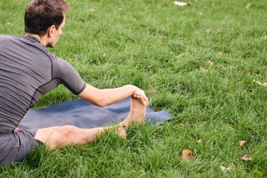 Spor giyimli genç bir adam parkta yoga yapıyor. Asana 'yı dışarıda pratik yap. İnsanlar yeşil çimlerde yoga minderiyle egzersiz yapıyorlar. Fitness ve sağlıklı yaşam tarzı. Sakinlik ve meditasyon kavramı.