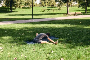 Spor giyimli genç bir adam parkta yoga yapıyor. Asana 'yı dışarıda pratik yap. İnsanlar yeşil çimlerde yoga minderiyle egzersiz yapıyorlar. Fitness ve sağlıklı yaşam tarzı. Sakinlik ve meditasyon kavramı.