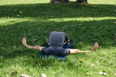 Spor giyimli genç bir adam parkta yoga yapıyor. Asana 'yı dışarıda pratik yap. İnsanlar yeşil çimlerde yoga minderiyle egzersiz yapıyorlar. Fitness ve sağlıklı yaşam tarzı. Sakinlik ve meditasyon kavramı.