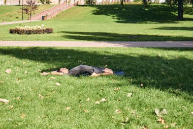 Spor giyimli genç bir adam parkta yoga yapıyor. Kapotasana asana 'yı dışarıda pratik yap. İnsanlar yeşil çimlerde yoga minderiyle egzersiz yapıyorlar. Fitness ve sağlıklı yaşam tarzı.