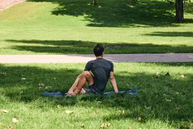 Spor giyimli genç bir adam parkta yoga yapıyor. Açık havada Omurga bükme egzersizi yap. İnsanlar yeşil çimlerde yoga minderiyle egzersiz yapıyorlar. Fitness ve sağlıklı yaşam tarzı.