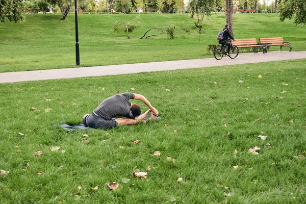 Spor giyimli genç bir adam parkta yoga yapıyor. Asana 'yı dışarıda pratik yap. Yeşil çimlerde yoga minderiyle egzersiz yapan insanlar. Parivritti Janu Sirshasana 'daki adam dizlerinin üzerine çöktü..