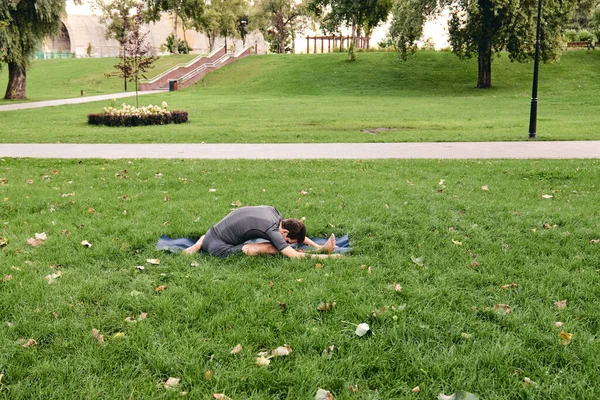 Spor giyimli genç bir adam parkta yoga yapıyor. Janu Sirshasana asana 'nın dizinden dizine kadar dışarıda poz ver. İnsanlar yeşil çimlerde yoga minderiyle egzersiz yapıyorlar. Fitness ve sağlıklı yaşam tarzı.