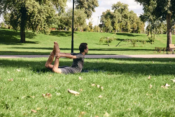 Spor giyimli genç bir adam parkta yoga yapıyor. Antrenman pruvası dışarıda asana pozu ver. İnsanlar yeşil çimlerde yoga minderiyle egzersiz yapıyorlar. Fitness ve sağlıklı yaşam tarzı.