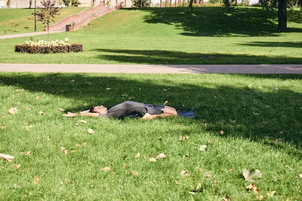 Spor giyimli genç bir adam parkta yoga yapıyor. Kapotasana asana 'yı dışarıda pratik yap. İnsanlar yeşil çimlerde yoga minderiyle egzersiz yapıyorlar. Fitness ve sağlıklı yaşam tarzı.