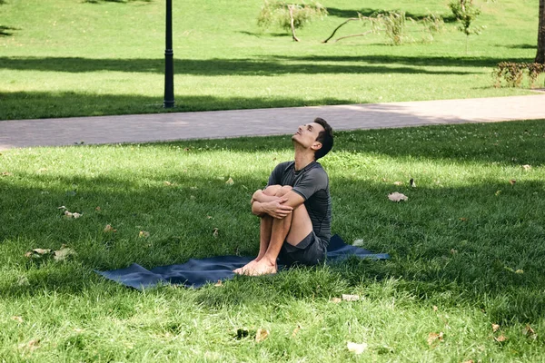 Spor giyimli genç bir adam parkta yoga yapıyor. Asana 'yı dışarıda pratik yap. İnsanlar yeşil çimlerde yoga minderiyle egzersiz yapıyorlar. Fitness ve sağlıklı yaşam tarzı. Sakinlik ve meditasyon kavramı.
