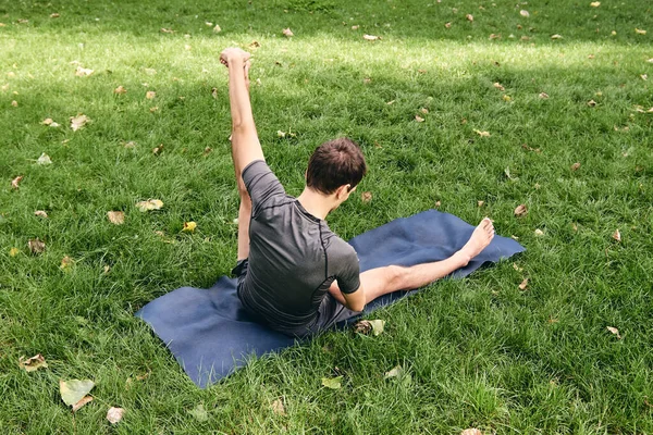 Spor giyimli genç bir adam parkta yoga yapıyor. Asana 'yı dışarıda pratik yap. İnsanlar yeşil çimlerde yoga minderiyle egzersiz yapıyorlar. Fitness ve sağlıklı yaşam tarzı. Sakinlik ve meditasyon kavramı.