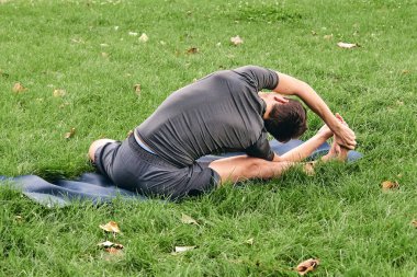 Spor giyimli genç bir adam parkta yoga yapıyor. Asana 'yı dışarıda pratik yap. Yeşil çimlerde yoga minderiyle egzersiz yapan insanlar. Parivritti Janu Sirshasana 'daki adam dizlerinin üzerine çöktü..
