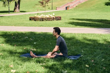 Spor giyimli genç bir adam parkta yoga yapıyor. Asana 'yı dışarıda pratik yap. İnsanlar yeşil çimlerde yoga minderiyle egzersiz yapıyorlar. Fitness ve sağlıklı yaşam tarzı. Sakinlik ve meditasyon kavramı.