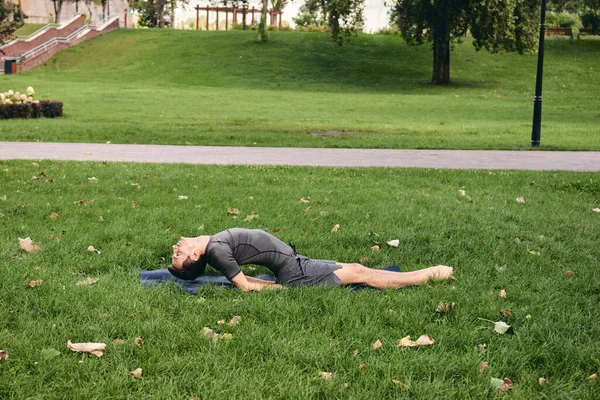 Spor giyimli genç bir adam parkta yoga yapıyor. Asana 'yı dışarıda pratik yap. İnsanlar yeşil çimlerde yoga minderiyle egzersiz yapıyorlar. Fitness ve sağlıklı yaşam tarzı. Sakinlik ve meditasyon kavramı.