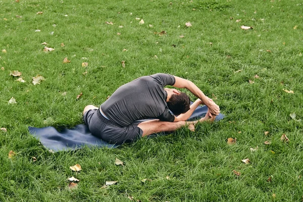 Spor giyimli genç bir adam parkta yoga yapıyor. Asana 'yı dışarıda pratik yap. Yeşil çimlerde yoga minderiyle egzersiz yapan insanlar. Parivritti Janu Sirshasana 'daki adam dizlerinin üzerine çöktü..
