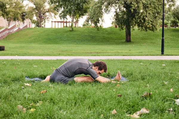 Spor giyimli genç bir adam parkta yoga yapıyor. Janu Sirshasana asana 'nın dizinden dizine kadar dışarıda poz ver. İnsanlar yeşil çimlerde yoga minderiyle egzersiz yapıyorlar. Fitness ve sağlıklı yaşam tarzı.