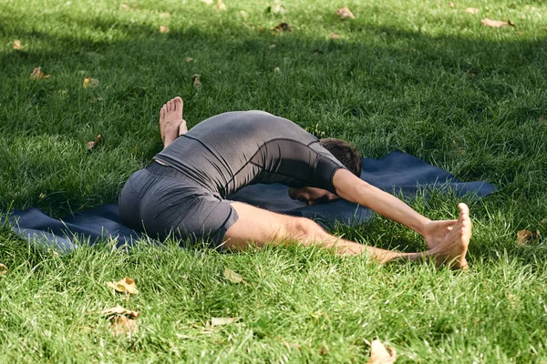 Young athletic man in sportswear doing yoga in the park. Practice asana outdoors. People exercising stretching on green grass with yoga mat. Fitness and healthy lifestyle. Concept of calm and meditation.