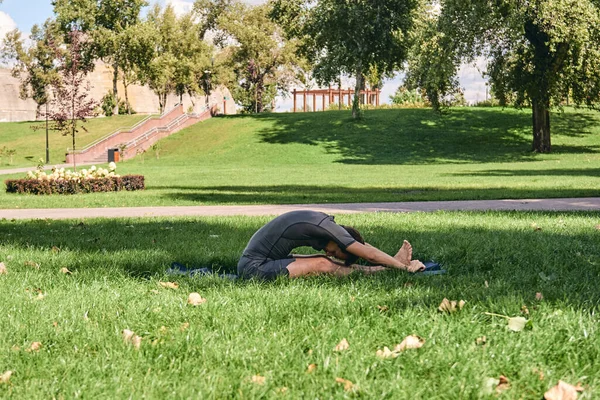 Spor giyimli genç bir adam parkta yoga yapıyor. Asana 'yı dışarıda pratik yap. İnsanlar yeşil çimlerde yoga minderiyle egzersiz yapıyorlar. Fitness ve sağlıklı yaşam tarzı. Sakinlik ve meditasyon kavramı.