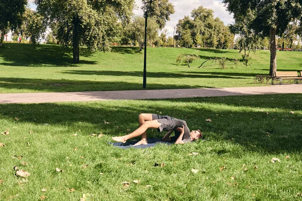 Spor giyimli genç bir adam parkta yoga yapıyor. Asana 'yı dışarıda pratik yap. Yeşil çimlerde yoga minderiyle egzersiz yapan insanlar. Balık pozu veren güçlü, olgun, beyaz adam..