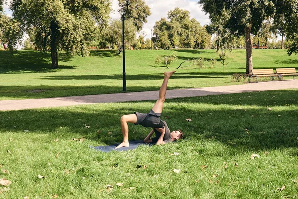 Young athletic man in sportswear doing yoga in the park. Practice asana poses outdoors. People exercising on green grass with yoga mat. Fitness and healthy lifestyle. Concept of calm and meditation.