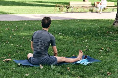 Spor giyimli genç bir adam parkta yoga yapıyor. Asana 'yı dışarıda pratik yap. İnsanlar yeşil çimlerde yoga minderiyle egzersiz yapıyorlar. Fitness ve sağlıklı yaşam tarzı. Sakinlik ve meditasyon kavramı.