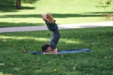 Spor giyimli genç bir adam parkta yoga yapıyor. Asana 'yı dışarıda pratik yap. Yeşil çimlerde yoga minderiyle egzersiz yapan insanlar. Nilüfer duruşu içinde zorlu amuda kalkmış güçlü beyaz adam..