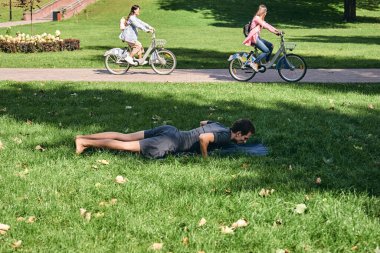 Spor giyimli genç bir adam parkta yoga yapıyor. Asana 'yı dışarıda pratik yap. İnsanlar yeşil çimlerde yoga minderiyle egzersiz yapıyorlar. Fitness ve sağlıklı yaşam tarzı. Sakinlik ve meditasyon kavramı.