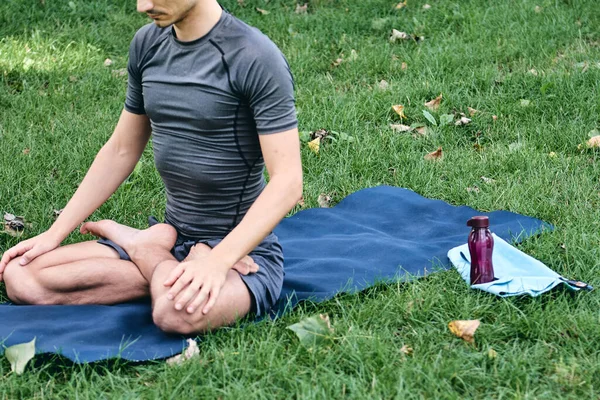 Spor kıyafetli genç adam parkta yoga yapıyor. Asana 'yı dışarıda pratik yap. Yeşil çimlerde yoga minderiyle egzersiz yapmak. Lotus pozunda oturan adam, sakin ve meditasyonlu. Fitness ve sağlıklı yaşam tarzı.