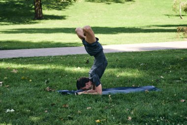 Spor giyimli genç bir adam parkta yoga yapıyor. Asana 'yı dışarıda pratik yap. Yeşil çimlerde yoga minderiyle egzersiz yapan insanlar. Nilüfer duruşu içinde zorlu amuda kalkmış güçlü beyaz adam..