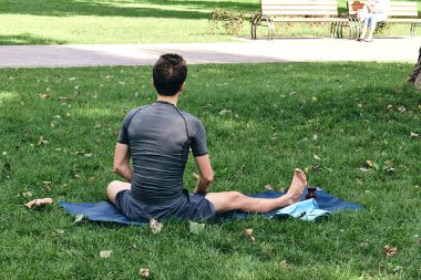 Spor giyimli genç bir adam parkta yoga yapıyor. Asana 'yı dışarıda pratik yap. İnsanlar yeşil çimlerde yoga minderiyle egzersiz yapıyorlar. Fitness ve sağlıklı yaşam tarzı. Sakinlik ve meditasyon kavramı.