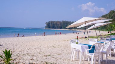 Şemsiyeler, masalar ve sandalyeler beyaz kumsalda içecekler, açık gökyüzünün altında mavi denizde kayıklar, güneşli bir günde insanlar yüzüyor.