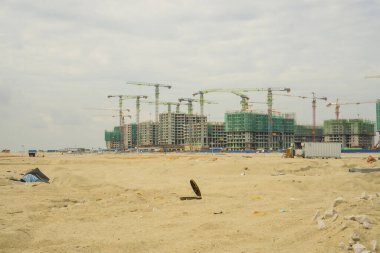 Büyük precast bina ve inşaat işinde uzun kule vinç yığınları, her hikaye üzerine iskeleler ve yeşil ağ korumasıyla kaplı, kirli gökyüzünün altındaki kumlu arazi, gelişim şehri ama aynı zamanda doğayı yok ediyor.