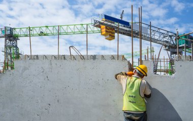 Bir inşaatçı binanın en üst katında çelik çubuklu beton yapı, turuncu güvenlik miğferi ve yeşil yelek, arka planda da bir vinç inşa ediyor.