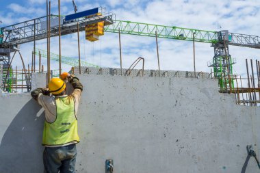 Bir inşaatçı binanın en üst katında çelik çubuklu beton yapı, turuncu güvenlik miğferi ve yeşil yelek, arka planda da bir vinç inşa ediyor.
