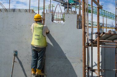 Bir inşaatçı binanın en üst katında çelik çubuklu beton yapı, turuncu güvenlik miğferi ve yeşil yelek, arka planda da bir vinç inşa ediyor.
