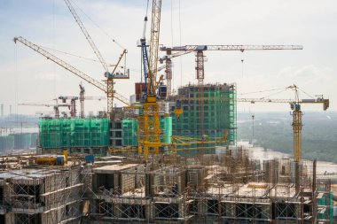 İnşaat alanında büyük bir binanın tepesinde birçok uzun vinç var. En üst katta hareket eden bir makine var. Her katta iskeleler ve yeşil ağ koruması var.