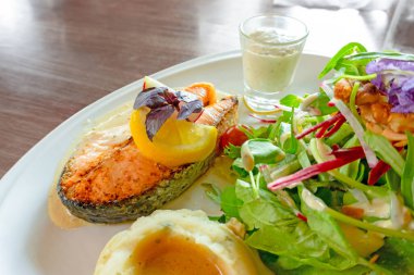 Izgara somon bifteği, beyaz tabakta dilimlenmiş, karışık sebze salatası, patates püresi ve bardakta sos, ahşap masada yemek ve kopyalama alanı.