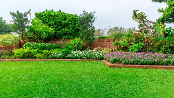 Güzel bir İngiliz kır evi bahçesi, pürüzsüz yeşil çimenler üzerinde renkli çiçekli bir bitki ve beyaz gökyüzünün altındaki halka açık bir parkın bakımını üstlenen bir grup yeşil ağaç. 