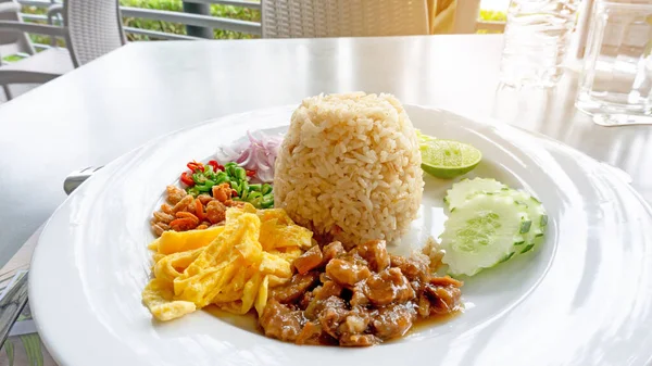 A dish of Thai food called Rice Mixed Shrimp Paste, the recipes are  fired rice, sweet pork, yellow omelet, dried shrimp, green and red hot chilli, onion, sliced cucumber and lemon in a white ceramic plate on gray table