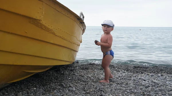 Küçük sevimli sevimli bebek çocuk tekne yakınında deniz kıyısında duruyor — Stok fotoğraf