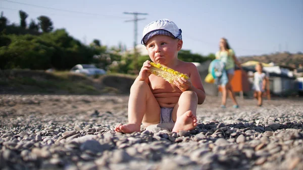 Little Cute Baby Boy siedzi na Kamyczki z brzegu i jeść kukurydza — Zdjęcie stockowe