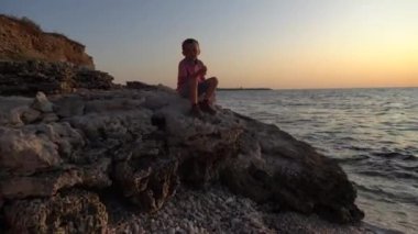 Denizde gün batımı bir arka plan karşı kaya üzerinde oturan küçük bir çocuğun silueti . Çocuk lezzetli bir şey yeme ve kameraya bakarak .
