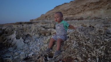 Gün batımında deniz kıyısındaki taşların üzerinde oturan çocuk. Çocuk kendi kendine konuşuyor ve kıpırdanıyor