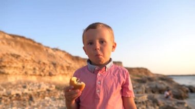 Yakın çekim, çocuk çocuk sahilde çörek yiyor. Gerçek olumlu duygular, gerçek zamanlı video görüntüleri, bir deniz kıyısında arka plan üzerinde çocuk, 4k