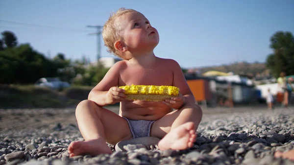 Niedliches Kind sitzt am Strand und hält Mais in den Händen und schaut in den Himmel — Stockfoto