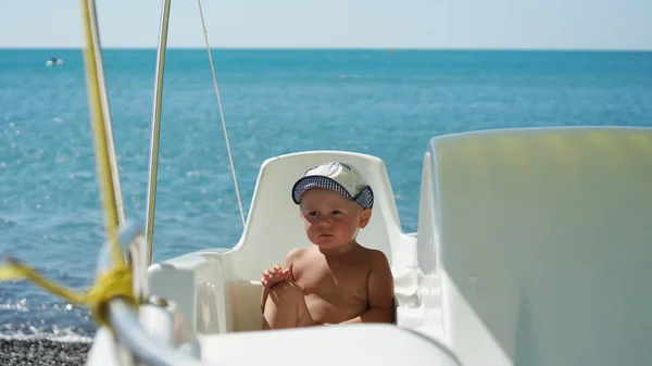 Chłopiec ubrany w kapelusz siedzący w łodzi na plaży. Dziecko na wakacjach na morzu — Zdjęcie stockowe
