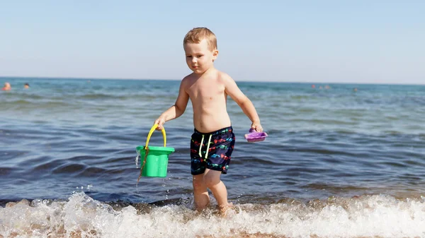 Mały chłopiec chodzący po morskiej plaży wzdłuż fali morskiej i trzyma wiadro w dłoniach — Zdjęcie stockowe