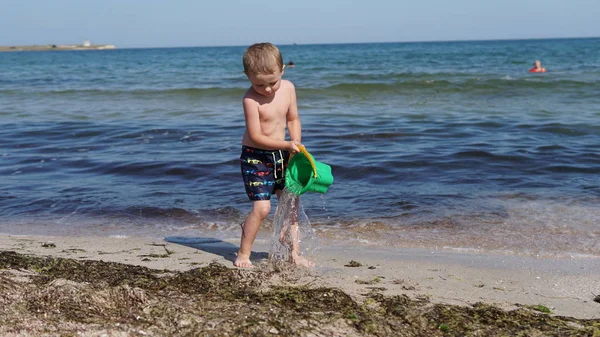 Mały chłopiec chodzenie na plaży morskiej i odlewania wody z wiadro w dłoniach — Zdjęcie stockowe