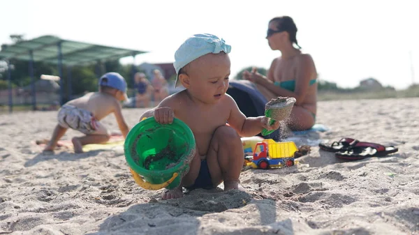 Criança agachada na praia do mar e brincando com areia com balde e colher . — Fotografia de Stock