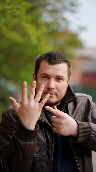 Portrait of a middle-aged bearded man in brown jacket on the sports field in the spring. Man looking in camera and pointing with finger to the other hand with fingers apart