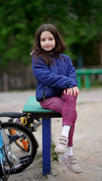 遊び場の公園で青いジャケットに長い緩い髪を持つかなり小さな女の子のサイクリストの肖像画 ベンチに座っている子供 — ストック写真