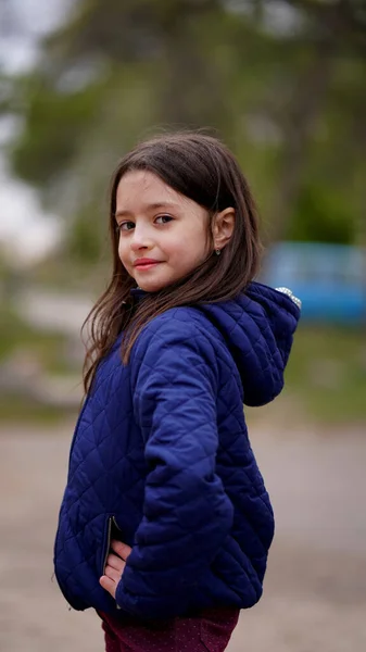 Parktaki parkta poz veren mavi ceketli, uzun sarı saçlı güzel bir kızın portresi. Gülümseyen çocuğun yan görünüşü