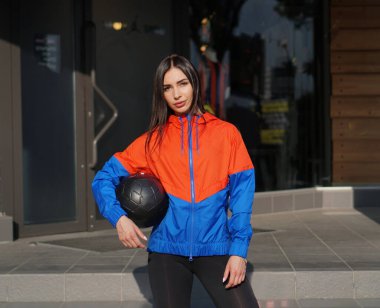 Uzun saçlı, kırmızı-mavi ceketli, mağazanın önünde topu olan çekici bir kadın. Modern spor modası kavramı.
